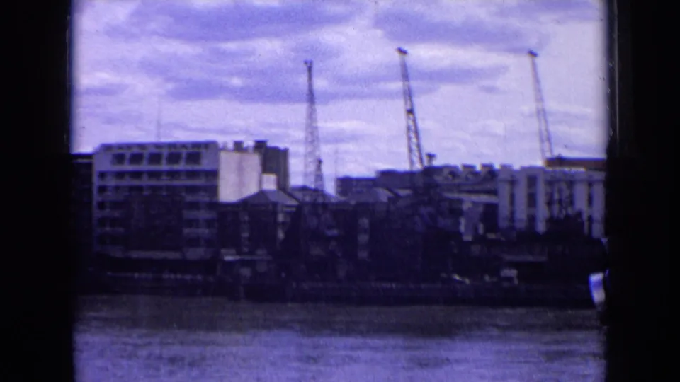 1963 England home-movie view across a river toward cranes and waterfront buildings, with dark vertical gate edges visible.