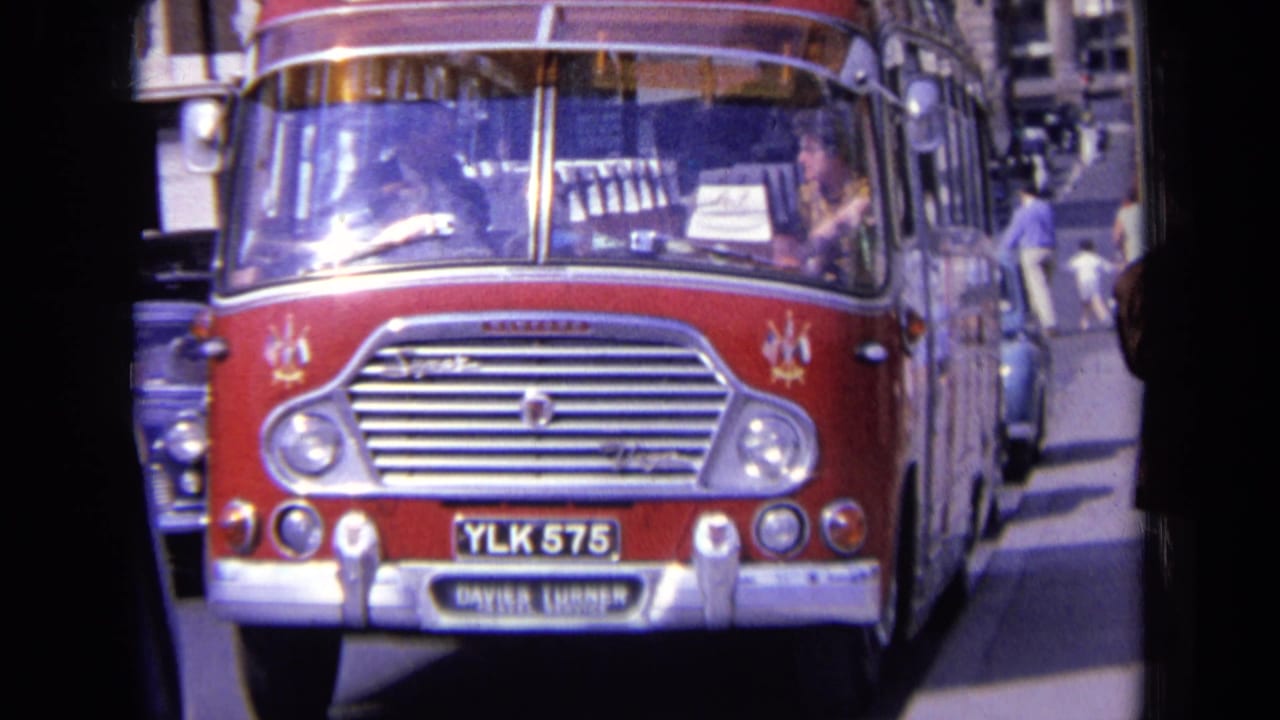 1963 England street frame showing a red bus with registration YLK 575, nearby cars, pedestrians, and bright windshield reflections.