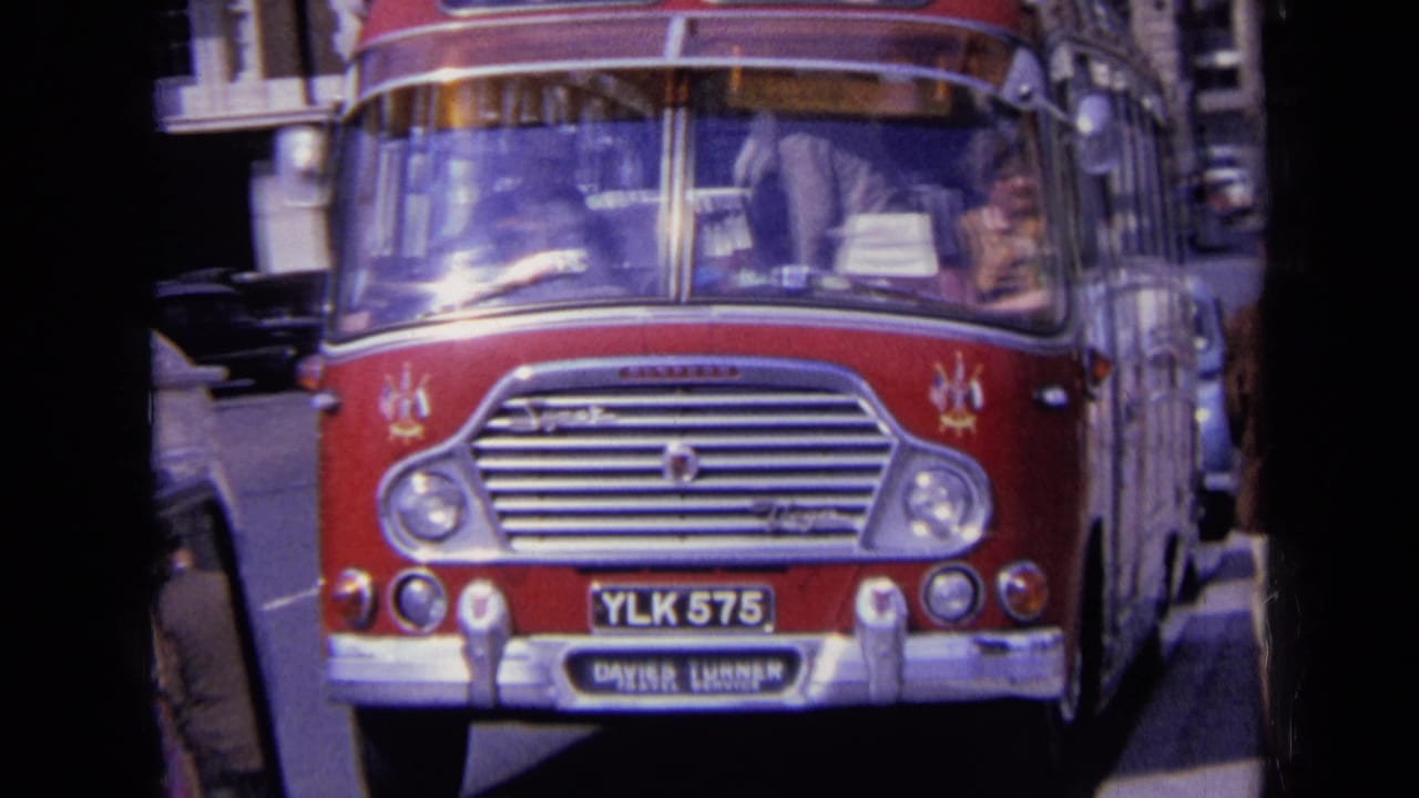 Red Daimler bus on an English city street, with the driver visible through reflective glass and period cars behind it.