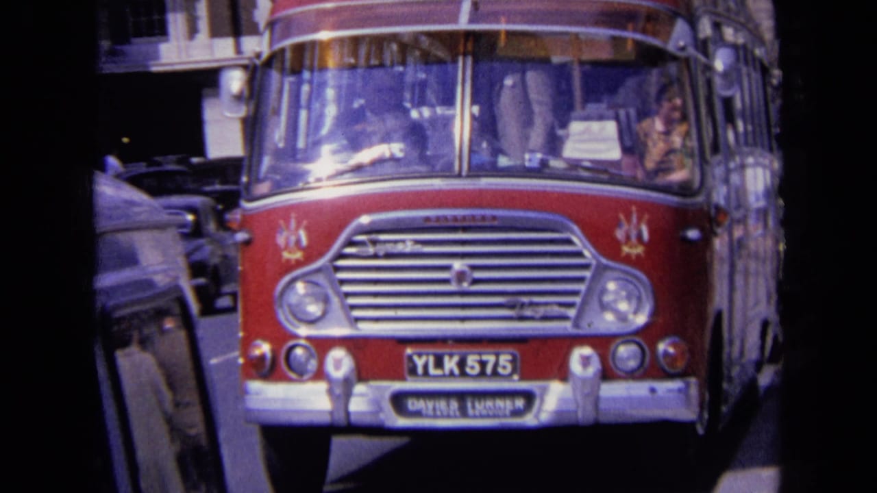 Close street-level 8mm view of a red Daimler bus in England, with YLK 575 plate, nearby car, and urban buildings partly visible.