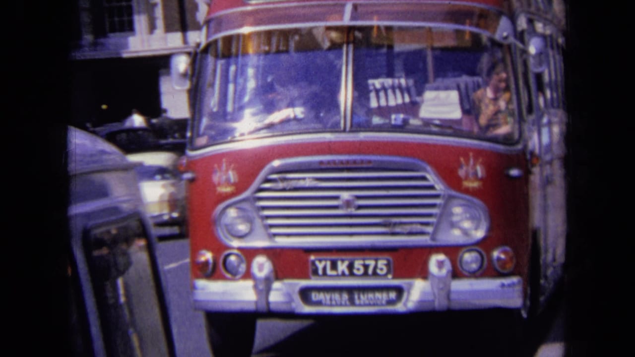 1960s England home-movie frame showing a red Daimler bus in traffic, with period cars, reflective windshield, and street buildings.