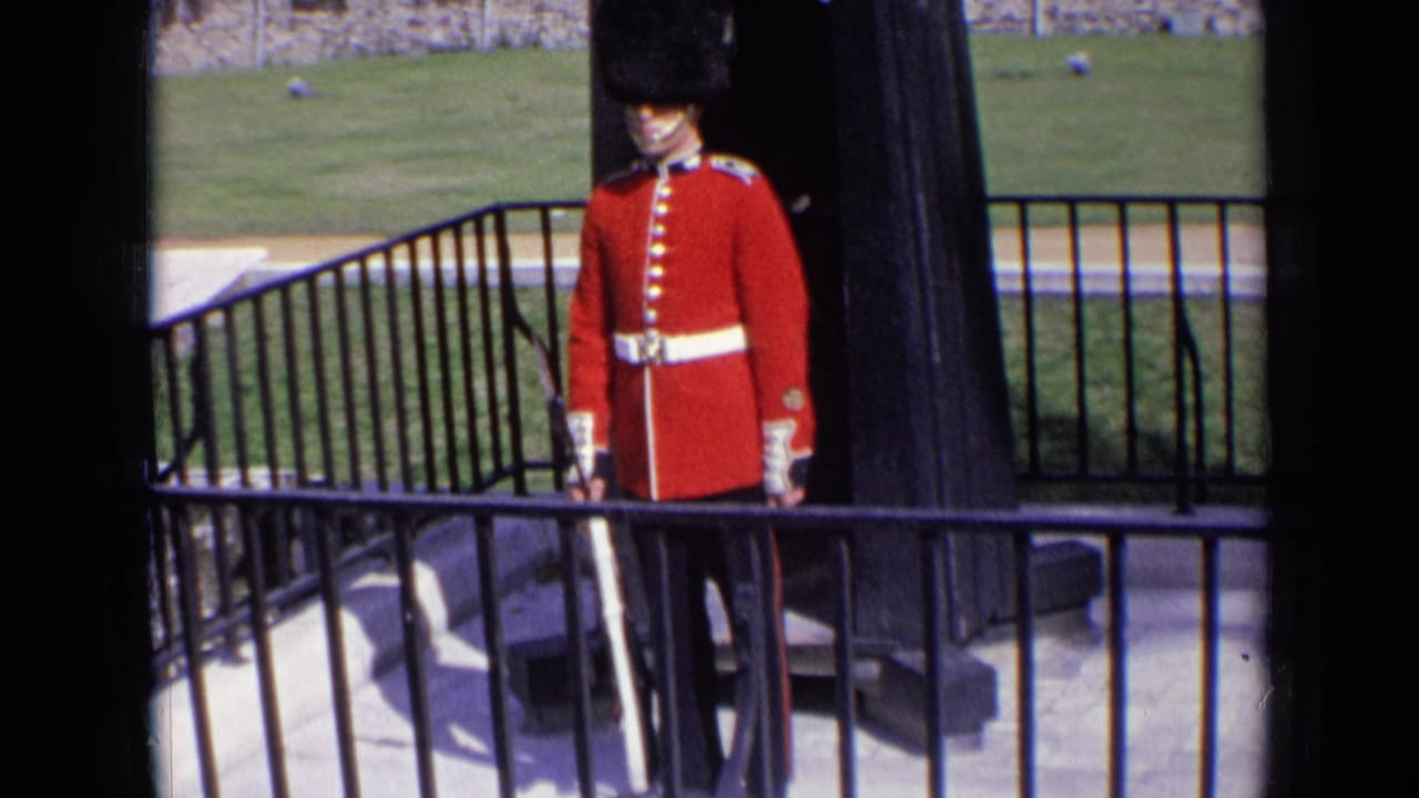 1963 England 8mm frame of a ceremonial guard in red tunic and tall black hat standing near railings and stone walls.