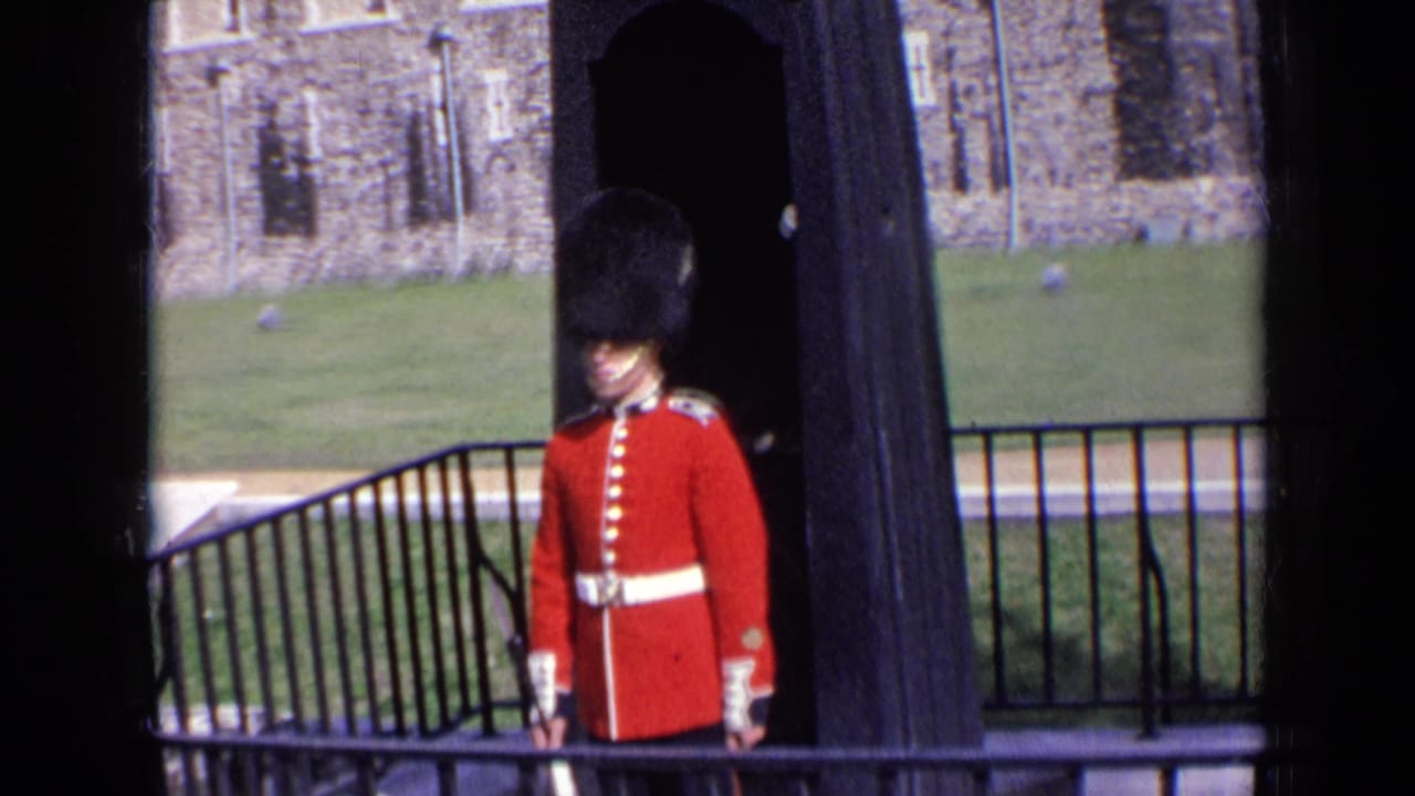 1963 England home-movie frame of a red-coated guard by a railing, with lawn and old stonework visible behind him.