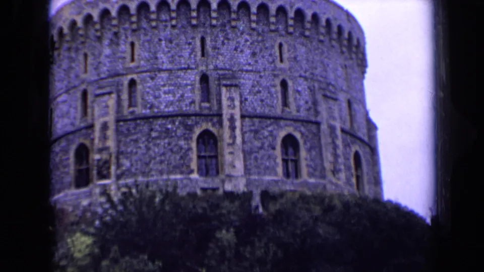 1963 England travel footage frame showing a round stone castle tower with arched windows above dark trees.