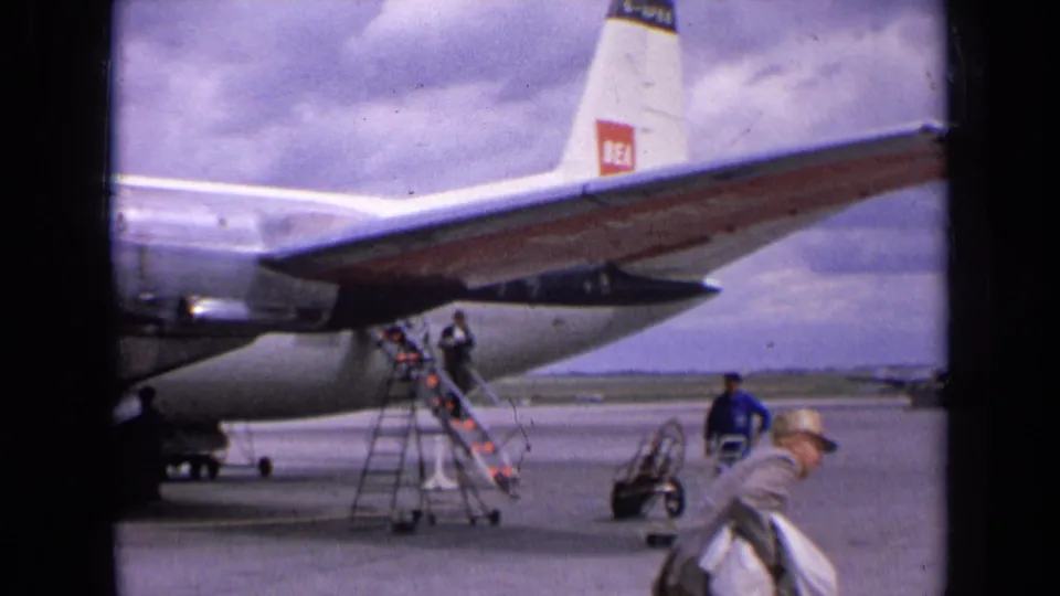 1963 England frame showing the rear of a BEA propeller plane, boarding stairs, ground equipment, and several people on the airport apron.