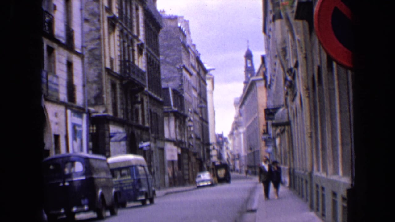 Narrow city street with parked vans, stone buildings and two pedestrians in the distance.