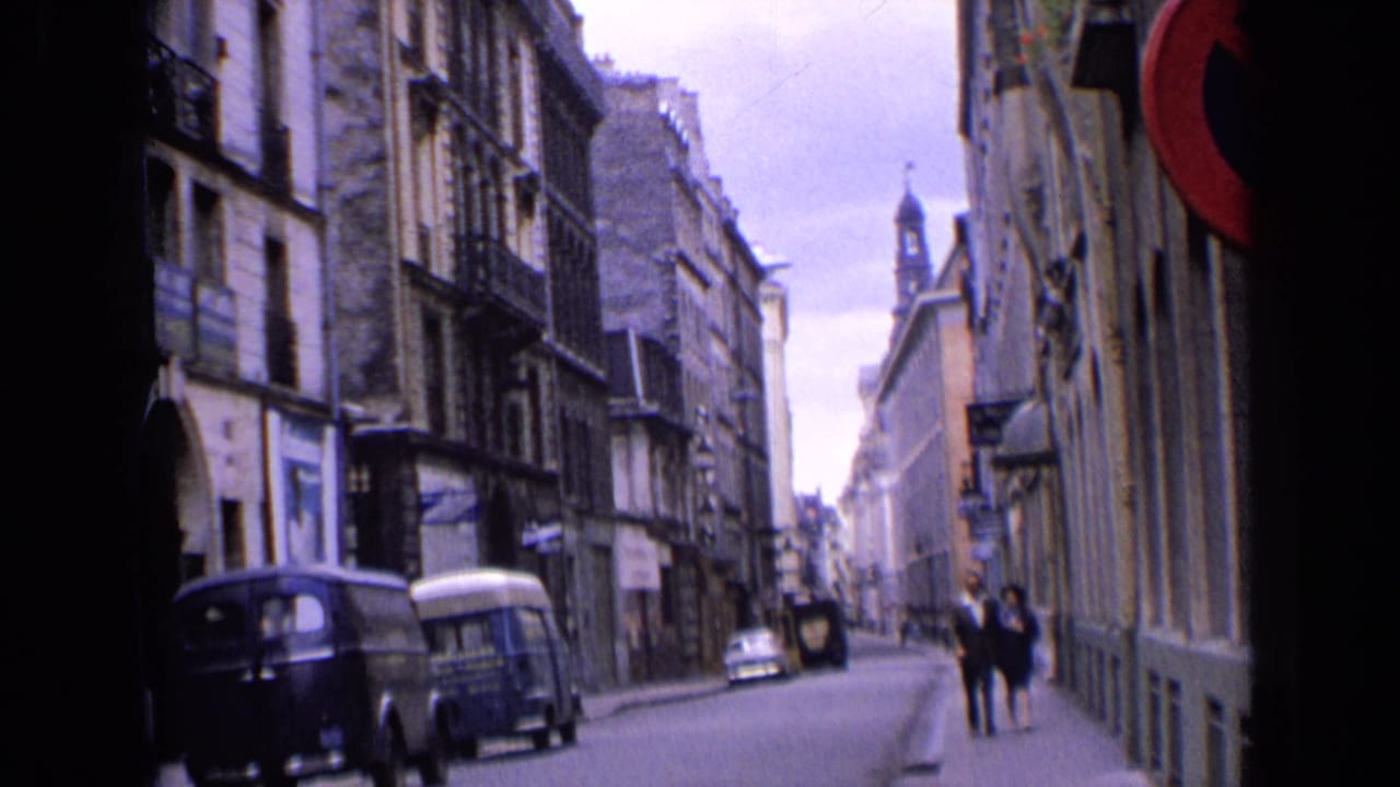 1963 England city street with a couple approaching the camera, older vans parked nearby and a spire above the rooftops.