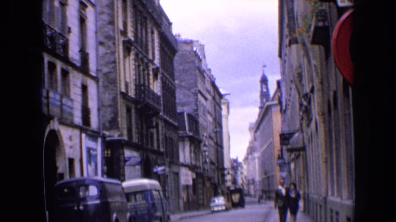 Home-movie view of a 1963 England street, showing a walking couple, parked vans and multi-story stone buildings.