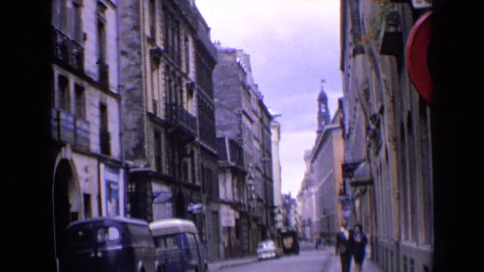 Home-movie view of a 1963 England street, showing a walking couple, parked vans and multi-story stone buildings.