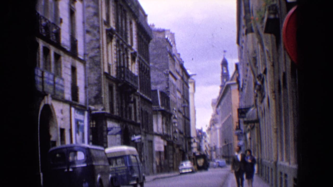 1963 England street footage with two adults walking toward the camera past parked vans and older urban architecture.
