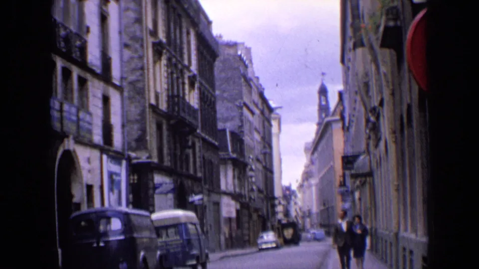 1963 England street footage with two adults walking toward the camera past parked vans and older urban architecture.