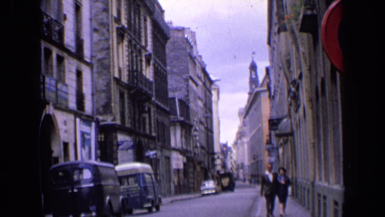 Narrow street with stone buildings, parked vans, pedestrians, and a distant spire.