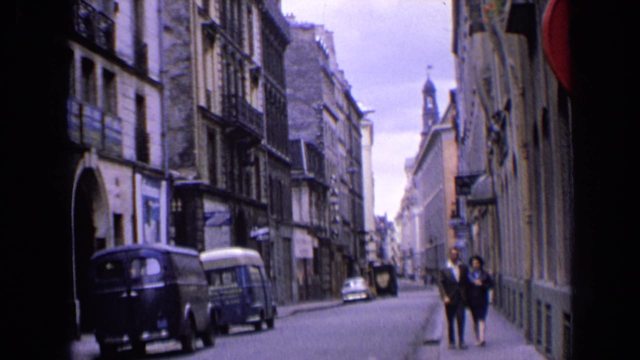 1963 England street scene showing parked vans, older stone buildings, a couple walking toward camera, and distant traffic.