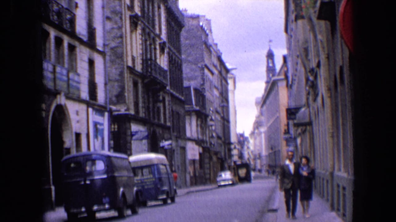 1963 England home-movie frame with a couple on the sidewalk, parked vans, old stone buildings, and a distant spire.