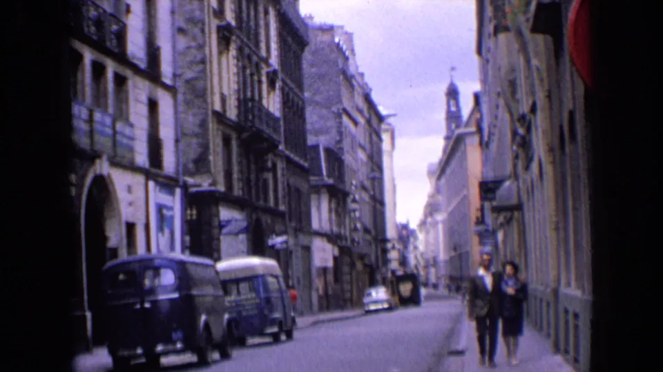 1963 England home-movie frame with a couple on the sidewalk, parked vans, old stone buildings, and a distant spire.