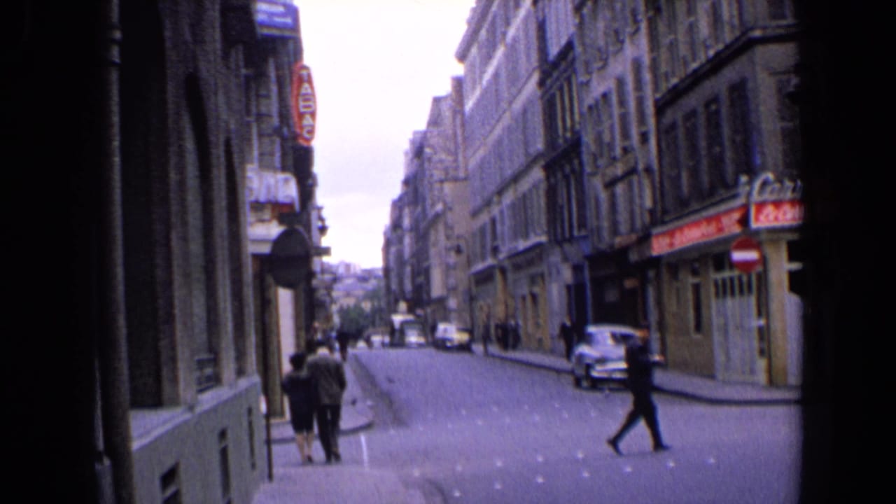 1963 England street view with pedestrians near a corner, shop signs, compact cars, and a suited man crossing the road.