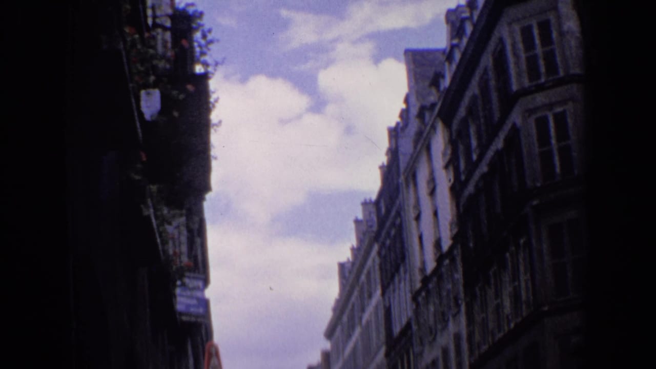 1963 England home-movie frame of a narrow urban street lined with older buildings beneath a cloudy sky.