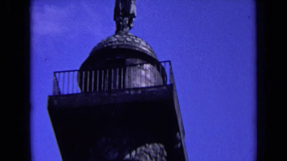 Handheld 8mm frame of a statue-topped monument, with railings and dark film edges visible against blue sky.