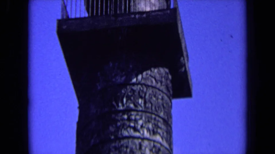 Low-angle 1963 England home-movie frame of an ornate monument column and platform beneath a statue against blue sky.