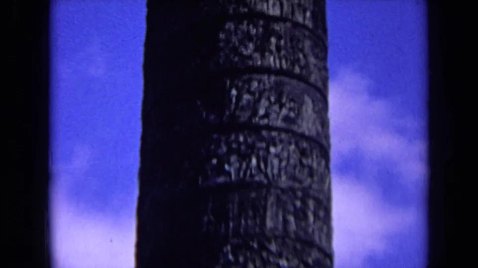 8mm frame showing the weathered column surface continuing downward, with pale clouds in the blue background.