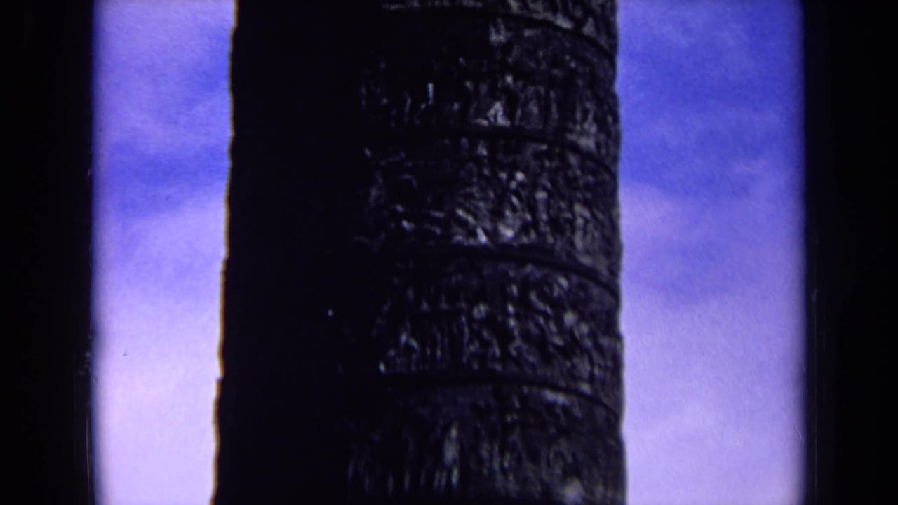 Dark carved column against a pale blue sky with black film edge at left.
