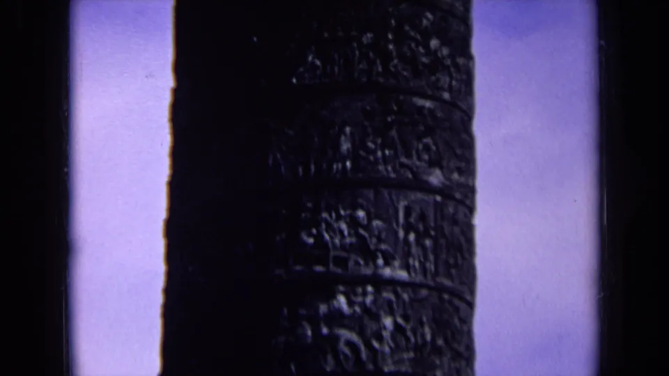 1963 England home-movie frame with a dark carved column rising beside a black vertical edge and blue sky.