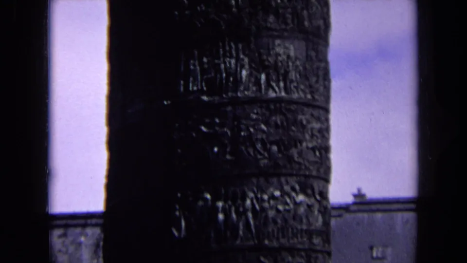 1963 England home-movie frame showing the carved column beside pale sky, with a small architectural roofline at the lower right.