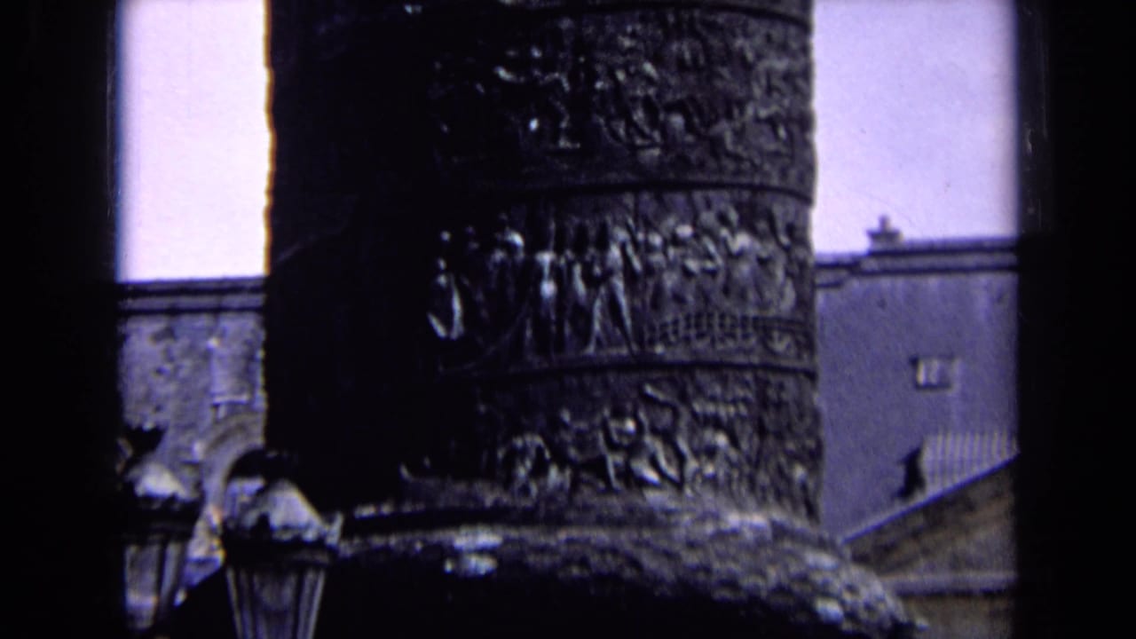 1963 England 8mm frame showing the upper section of a carved public monument with relief bands, lamps and building edges in the background.