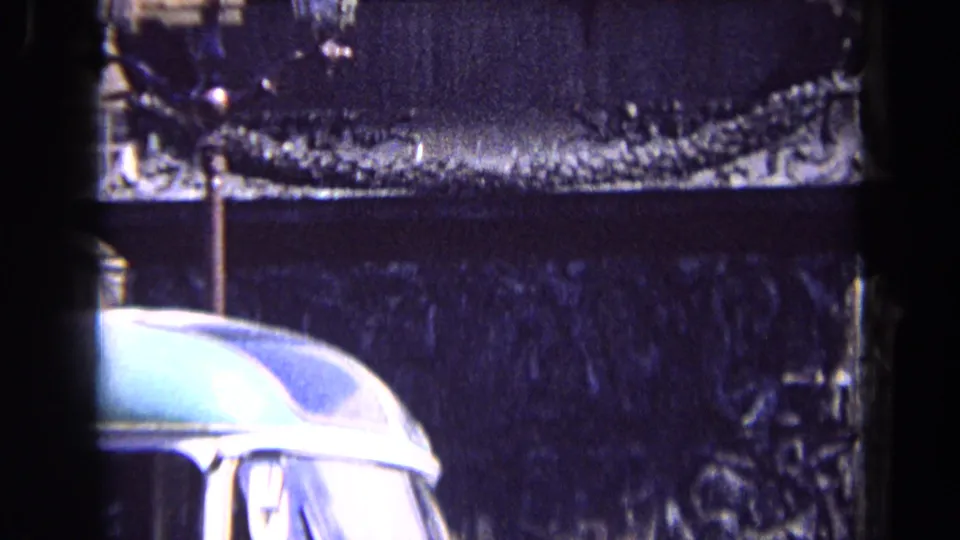 1963 England frame showing the monument base behind a rounded car roof, with street lamps and building edges partly visible.