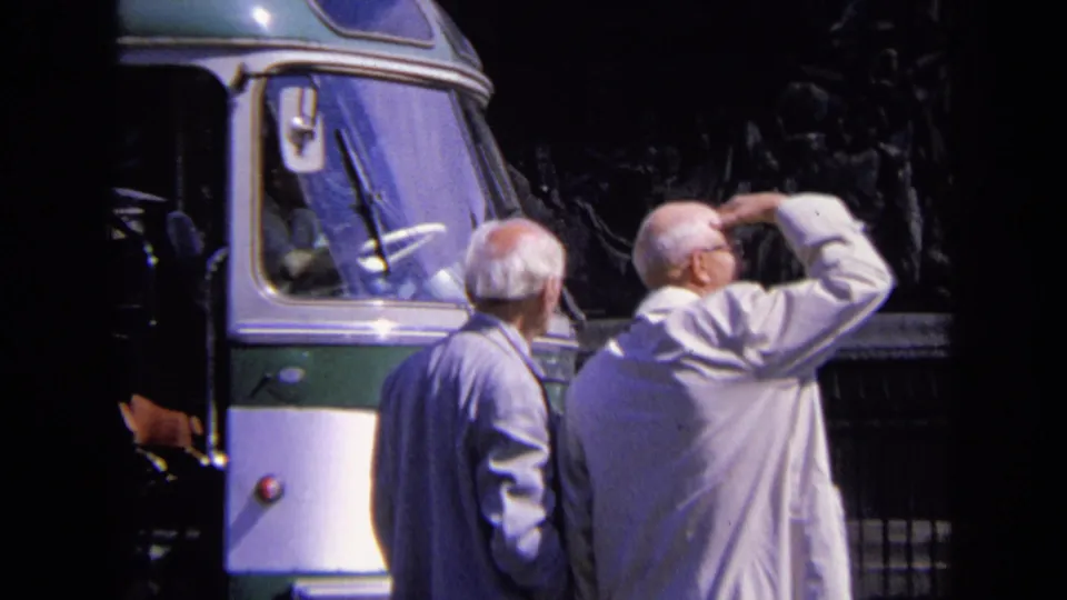 Two older men in light jackets stand beside a bus in Paris, one raising his hand as if shading his eyes.