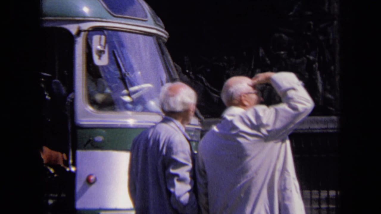 Two older men stand near a green-and-white bus beside the Arc de Triomphe.
