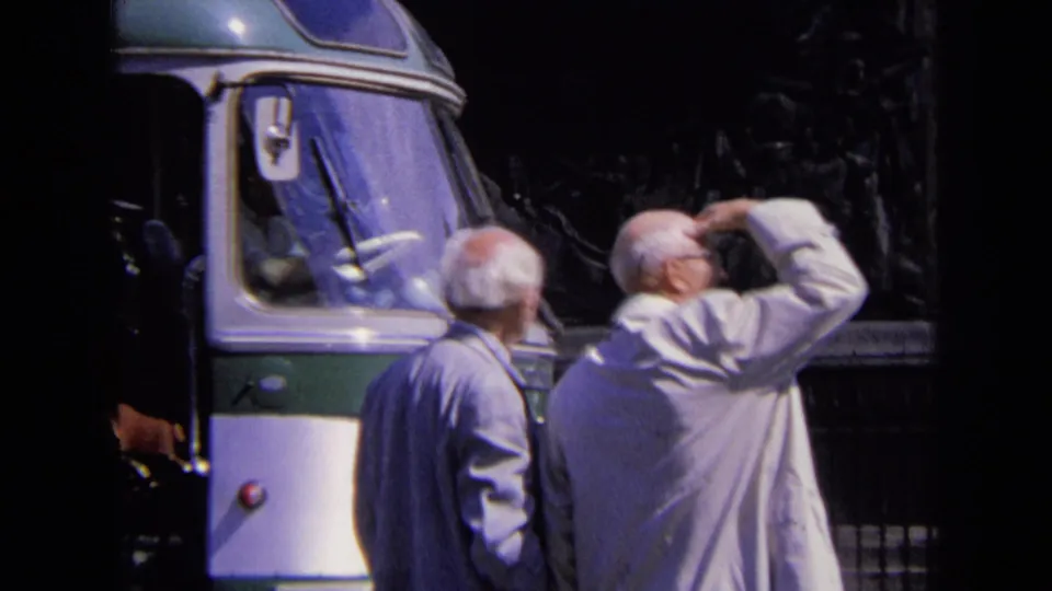 1963 Paris home-movie frame showing two older men beside a green-and-white bus near the Arc de Triomphe.