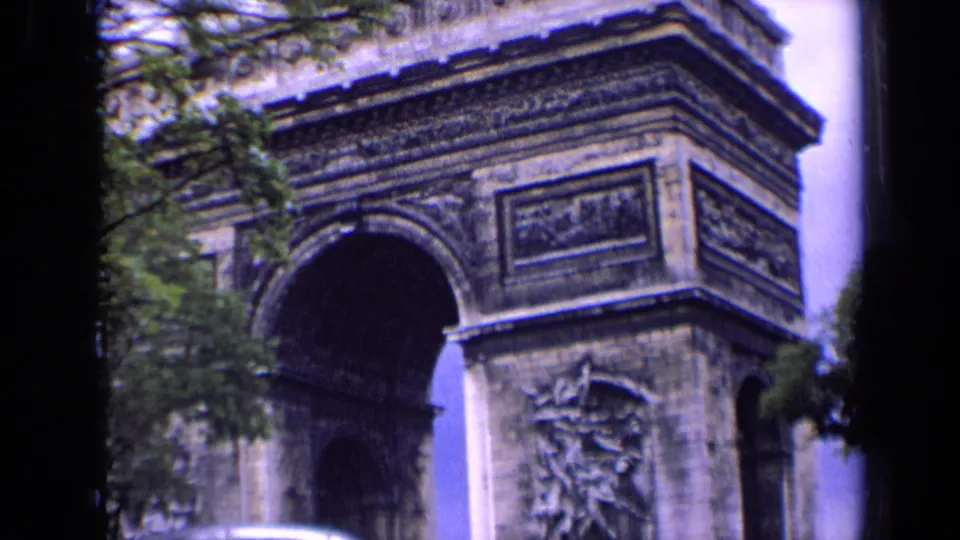 Tight 1963 Paris home-movie view of the Arc de Triomphe with sculpted panels, trees, and a bus edge below.