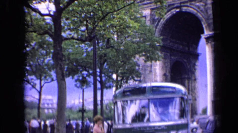 A green-and-white bus passes near the Arc de Triomphe in 1963 Paris, with pedestrians visible in the background.