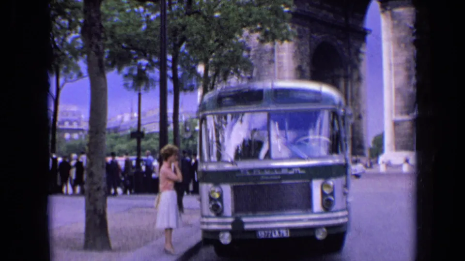 1963 Paris home-movie frame with a bus by the Arc de Triomphe and a woman in a light skirt standing on the sidewalk.