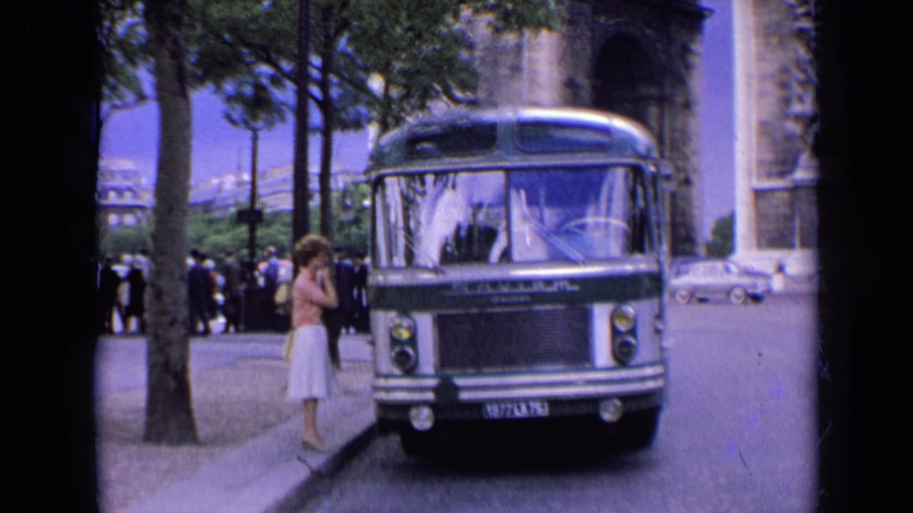 1963 France home-movie frame showing a parked bus, a woman at the curb, pedestrians, trees, and the Arc de Triomphe.