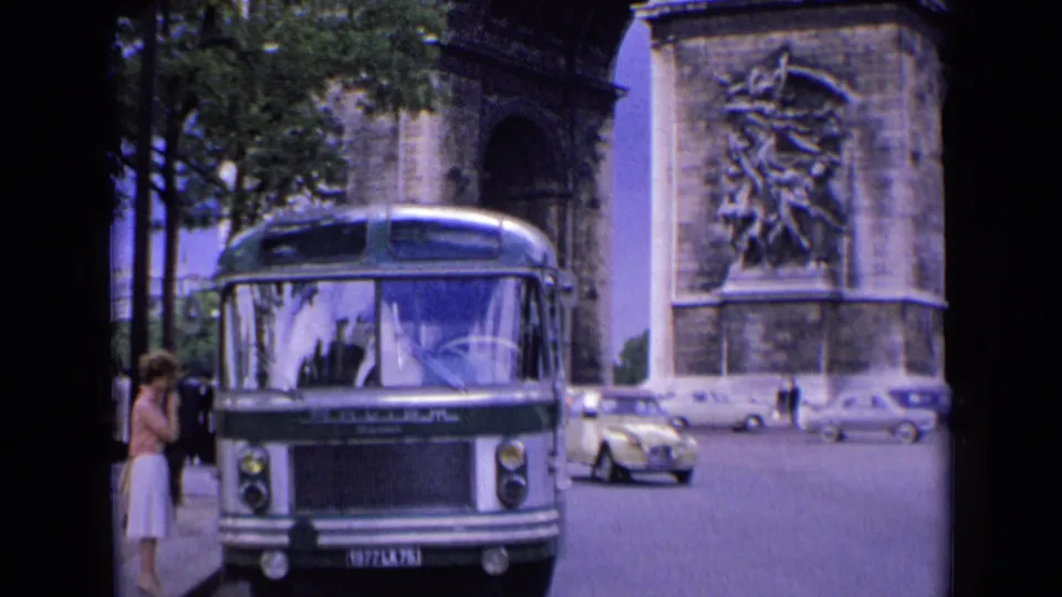 1963 Paris street view with a green-and-white bus, woman at the curb, passing cars, and the Arc de Triomphe behind.