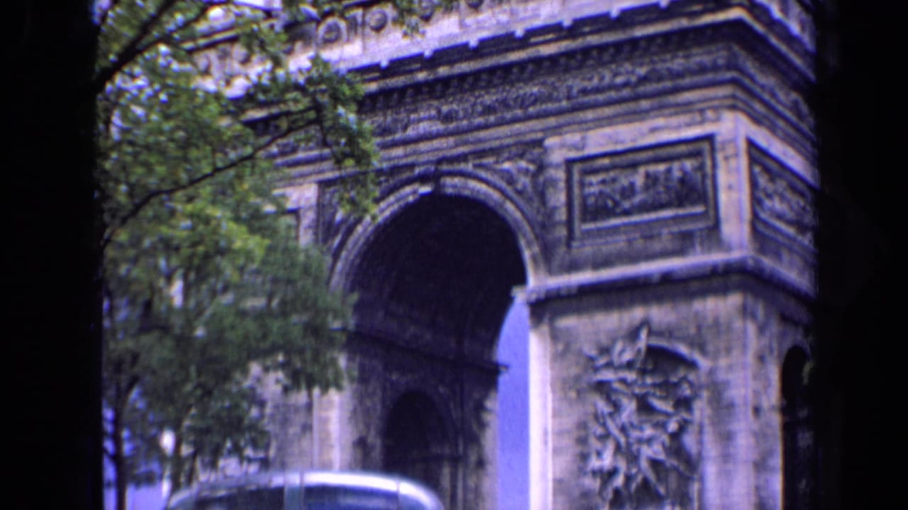 1963 Paris home-movie frame showing the Arc de Triomphe from a visitor viewpoint, with trees, street edge, and soft 8mm color.