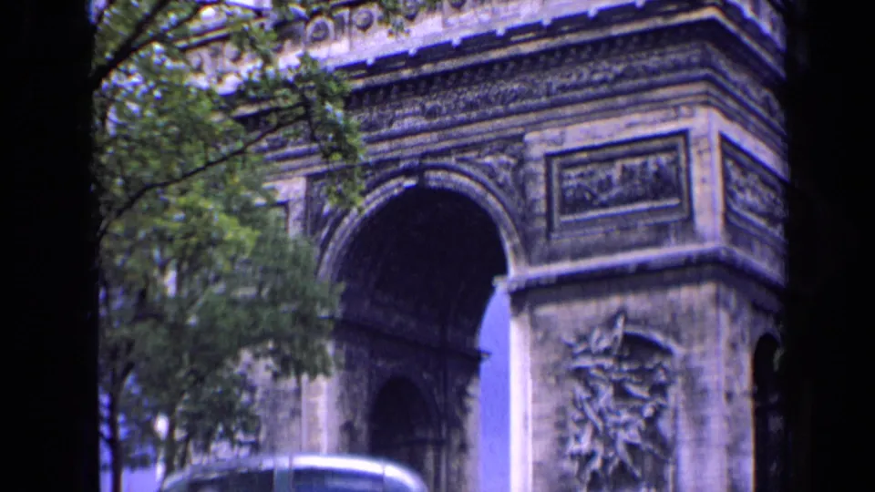 1963 Paris home-movie frame showing the Arc de Triomphe from a visitor viewpoint, with trees, street edge, and soft 8mm color.