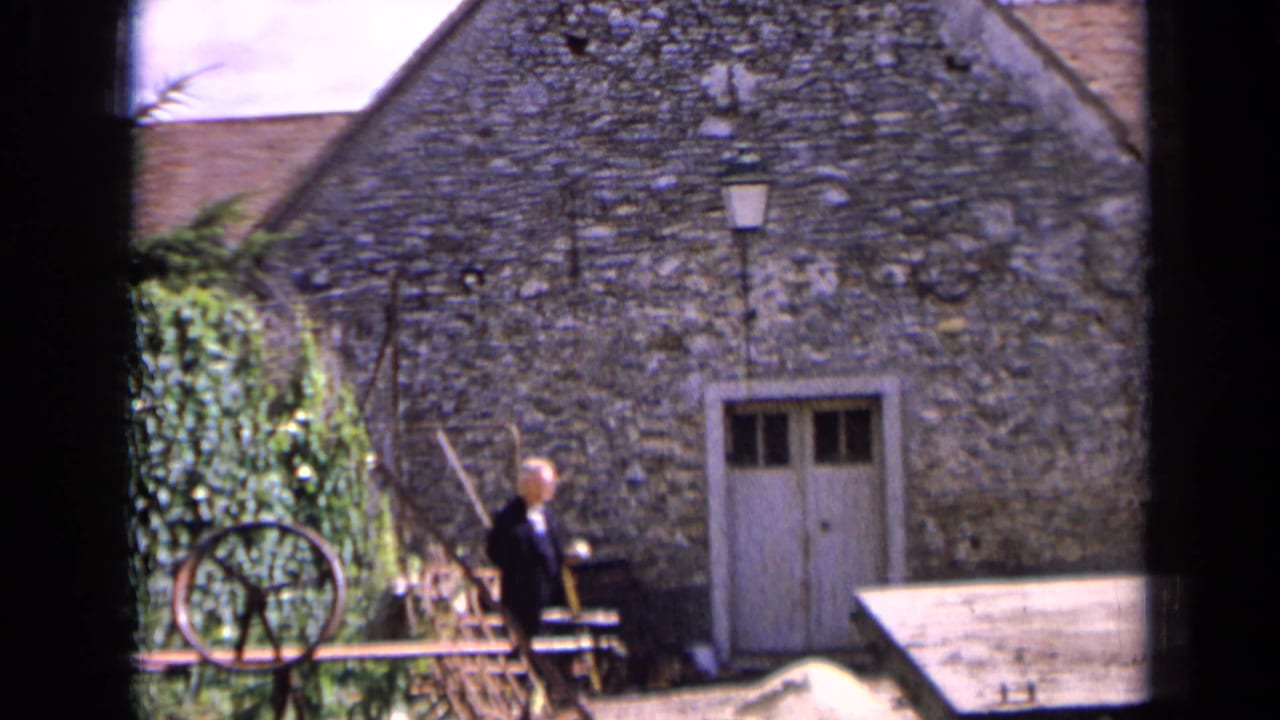 1963 France home-movie frame showing a man walking past a rough stone building with a pale door, lantern, and wheel at the edge.