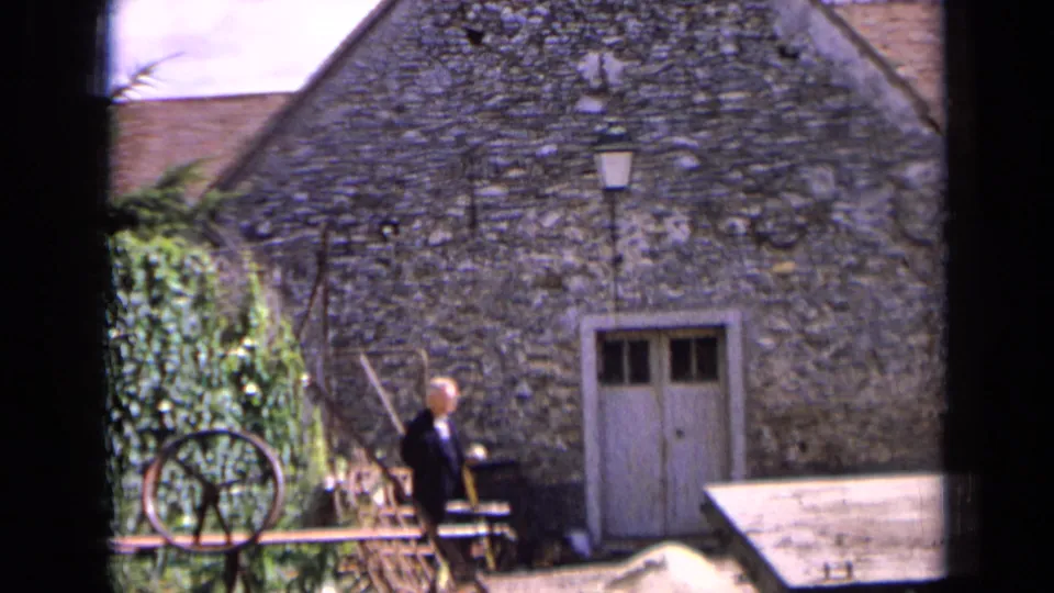 1963 France home-movie frame showing a man walking past a rough stone building with a pale door, lantern, and wheel at the edge.