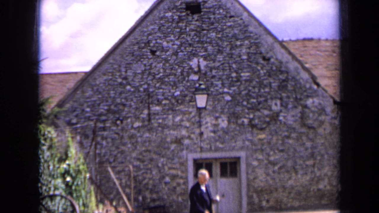 1963 France frame with a small figure in dark clothing moving past a textured stone wall, pale doorway, and mounted lantern.