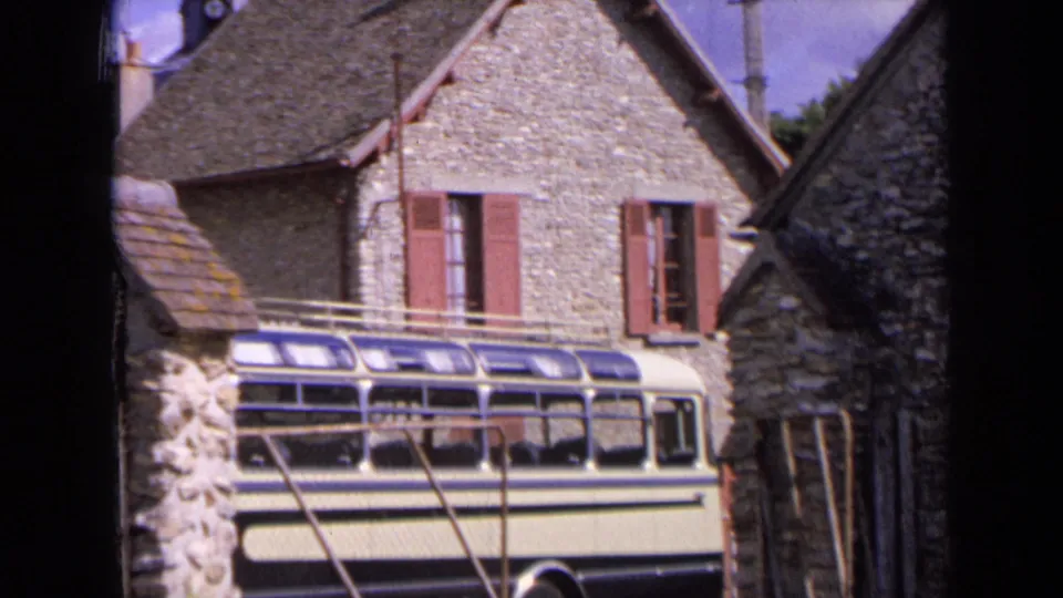 1963 France home-movie frame showing a bus beside stone village buildings with red shutters, rooflines, and bright sky.