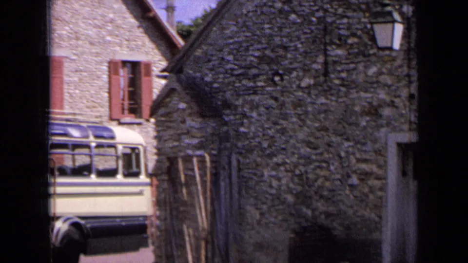 1963 France frame showing the rear portion of a bus near rustic stone walls, red shutters, a lantern, and a pale door.