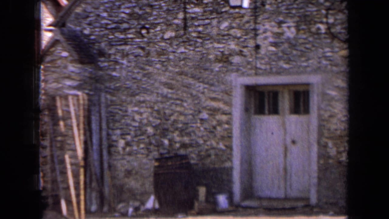 1963 France home-movie frame showing a rustic stone facade with a white wooden door and a dark barrel beside the entrance.