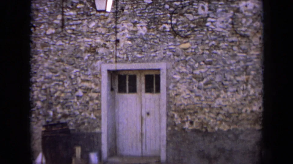1963 France home-movie frame holding on the weathered white door within a rough stone wall, with a barrel just to the left.