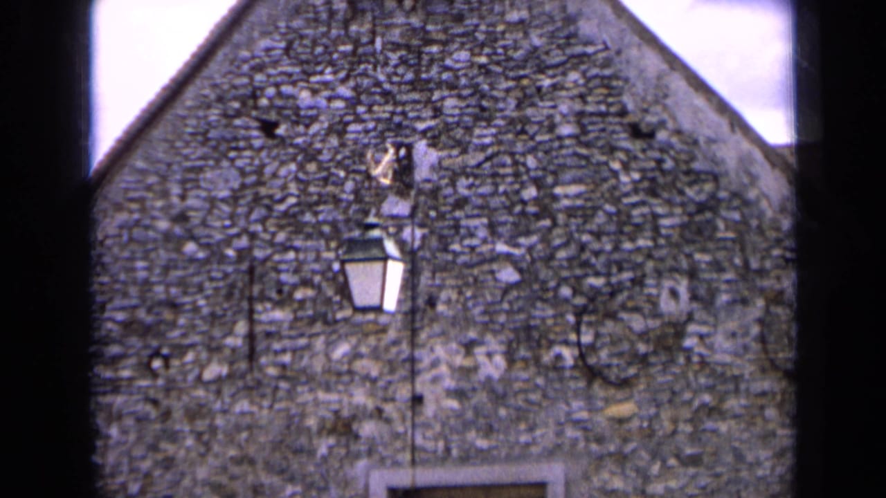 1963 France home-movie frame showing a rough stone gabled building, small window opening, and wall-mounted lantern in sun.