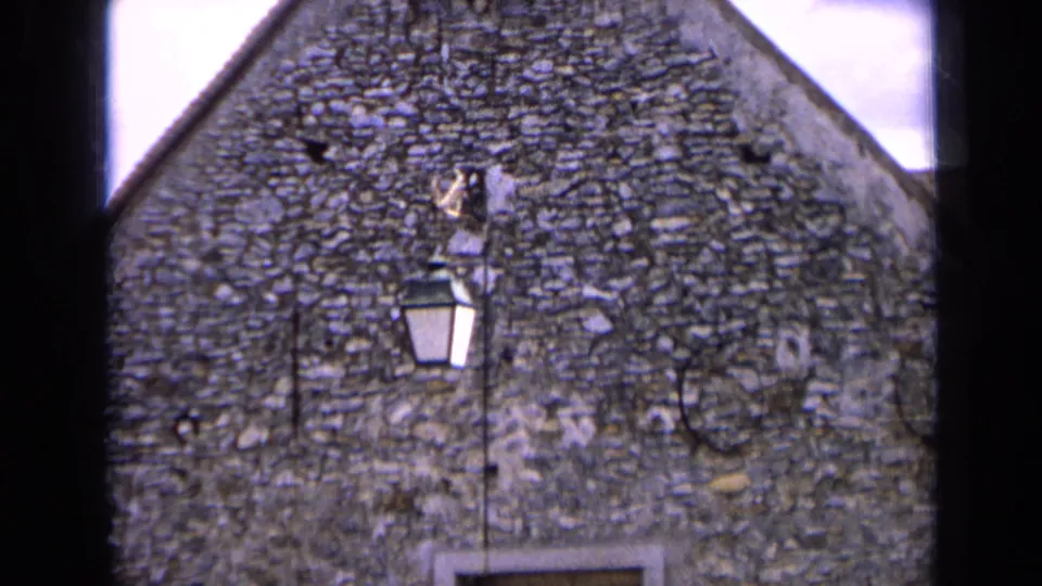 1963 France home-movie frame showing a rough stone gabled building, small window opening, and wall-mounted lantern in sun.