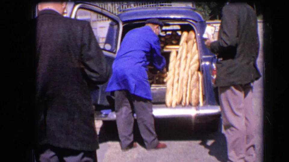 A man in a blue coat places long baguettes into an open car boot as two adults stand nearby in 1963 France.