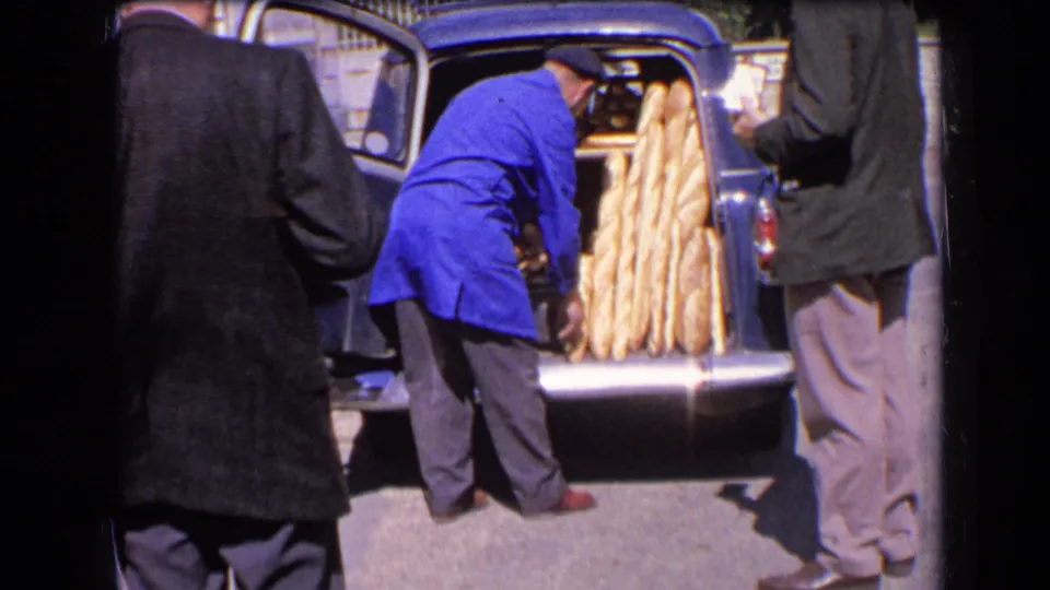The bread-loading continues at the rear of a blue vintage car, with adults gathered around the open boot in 1963 France.